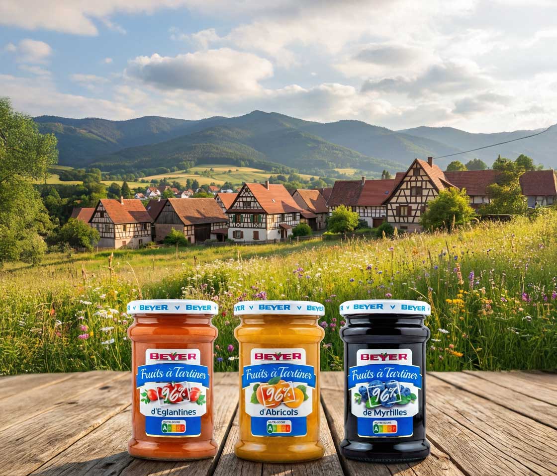 Deux pots de la gamme Beyer "Les 100%" (Cerises Noires et Rhubarbe) pos�s sur une table en bois rustique. L'arri�re-plan montre un village alsacien pittoresque nich� dans une vall�e verdoyante au pied de montagnes escarp�es.